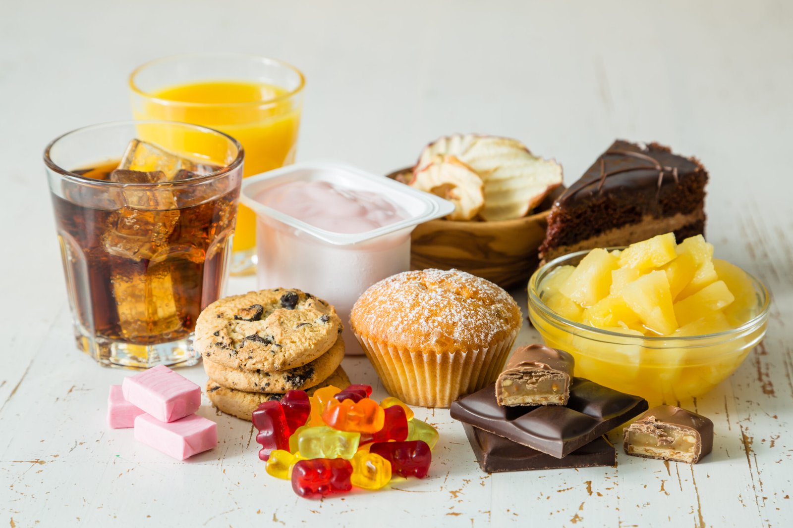 Selection of sugary foods and drinks including soda, fruit juice, yogurt, cookies, cake, sweets, chocolate, muffins, and canned pineapple displayed on a table to illustrate common dietary sources of sugar and processed foods